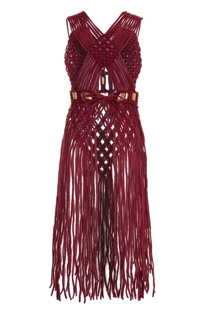 A handcrafted macramé dress in a deep burgundy color, featuring intricate knotted patterns, thin shoulder straps, and gold accents. The dress has a fitted bodice with detailed weaving, and long, fringed strands cascading down from the waist, creating a boho-chic aesthetic