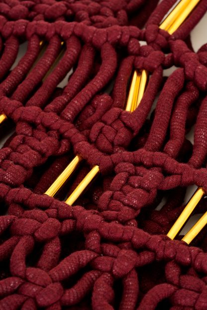 Close-up of handcrafted macramé dress in a deep burgundy color, featuring intricate knotted patterns, thin shoulder straps, and gold accents. The dress has a fitted bodice with detailed weaving, and long, fringed strands cascading down from the waist, creating a boho-chic aesthetic