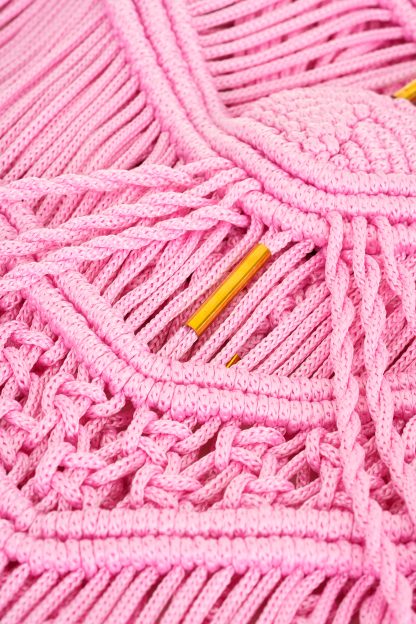 Close-up of a pink macramé fabric with intricate knotting details and fringe, featuring gold metal accents woven into the design. The textured pattern highlights the craftsmanship of the woven threads.